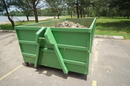 A large, rectangular green skip with reinforced metal sides and a smooth, glossy finish, positioned on a paved parking area near a grassy park or garden. The skip is filled with a mixture of garden waste and debris, including twigs, leaves, and plant material, which are visible over the top edge. In the background, there are mature trees with leafy branches, a lake or pond, and a continuation of the green lawn. The skip is slightly to the right of the image, with a small gap visible on its left side, and is situated on an area marked with yellow lines. The scene is illuminated by natural daylight, suggesting a bright, clear day suitable for garden clearance or rubbish removal activities. This image illustrates typical waste management equipment used by companies such as Garden Clearance Dulwich in providing garden clearance services, emphasizing the type of rubbish handled and the environment in which such containers are placed.