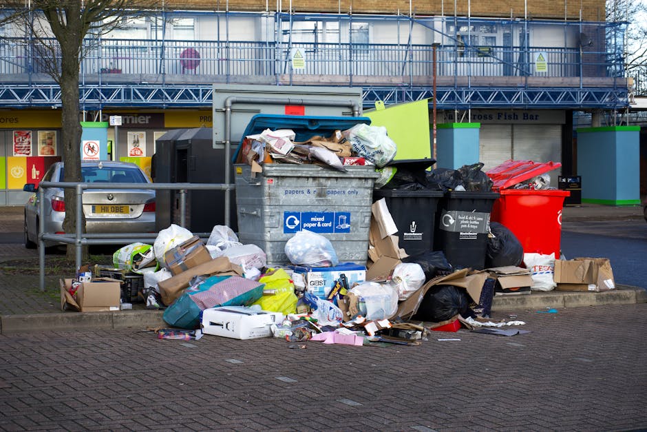 A sanitation worker wearing a yellow high-visibility vest and black trousers is engaged in rubbish collection in an urban area, standing next to a large red waste collection vehicle with the back open