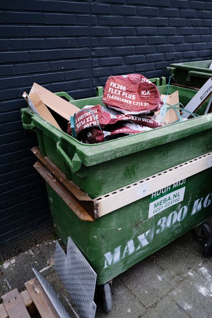 Two large black plastic refuse bags filled with waste are placed on the pavement near the curb in an outdoor urban setting, with one bag positioned upright and the other leaning against a wooden fence